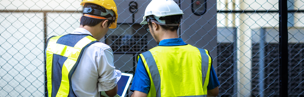 Two workers look at a laptop screen in front of some switchgear